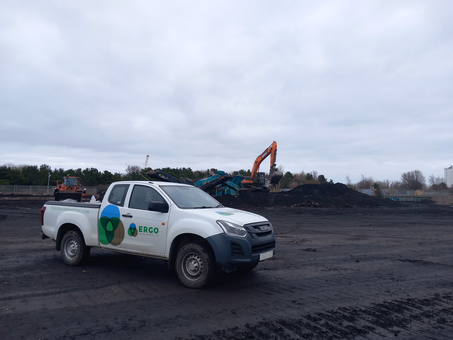 Parked white Suzuki ISUZU truck from ERGO Environmental on brownfield soil with various diggers and heap of soil in the background